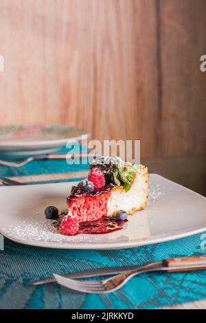 Gustosa cheesecake con marmellata rossa, lamponi maturi, mirtilli, foglie di menta fresca e zucchero a velo in piatto bianco di ceramica nel ristorante Foto Stock