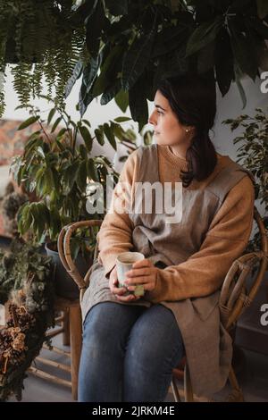 Donna rilassata e penosa in abiti casual guardando lontano e contemplando mentre si siede sul divano accanto a un piccolo tavolo di legno e bere gustoso caffè agai Foto Stock