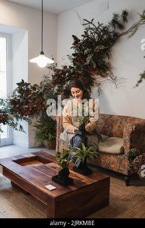 Angolo alto di gioioso fiorista godendo il proprio risultato di lavoro mentre si siede sul divano accanto a un piccolo tavolo di legno e tenendo verde pianta in vaso contro l'interno Foto Stock