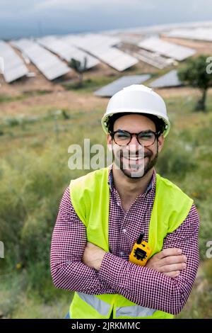 Felice ingegnere maschio con le braccia incrociate sorridendo guardando la macchina fotografica mentre si trova in piedi su sfondo sfocato di pannelli fotovoltaici e cielo nuvoloso Foto Stock