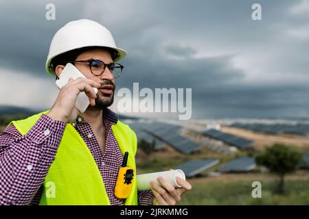 Uomo in elmetto ai pannelli fotovoltaici mentre si discute del lavoro di centrale solare su smartphone Foto Stock