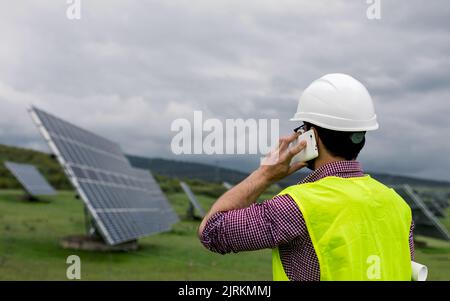 Vista posteriore dell'uomo anonimo in hardhat ai pannelli fotovoltaici mentre si discute del lavoro di centrale solare su smartphone Foto Stock