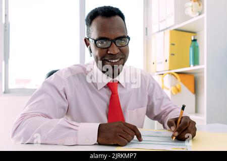Maturo afroamericano maschio executive con auricolare in camicia bianca con cravatta e occhiali prendendo nota su documento di carta a tavola nello spazio di lavoro Foto Stock
