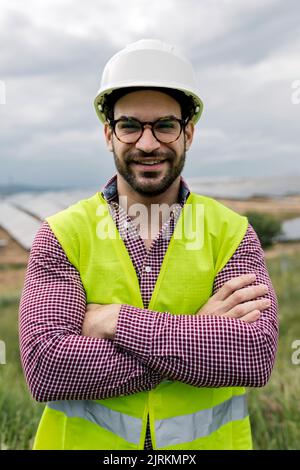 Felice ingegnere maschio con le braccia incrociate sorridendo guardando la macchina fotografica mentre si trova in piedi su sfondo sfocato di pannelli fotovoltaici e cielo nuvoloso Foto Stock