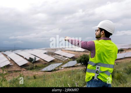 Uomo in elmetto che indica i pannelli fotovoltaici mentre lavora sul sito della centrale di energia solare sullo smartphone Foto Stock