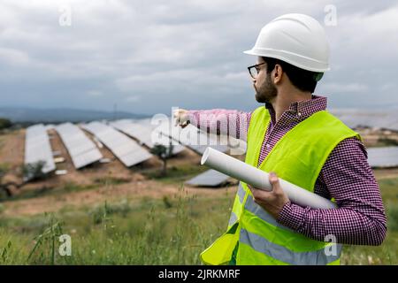 Uomo in elmetto che indica i pannelli fotovoltaici mentre lavora sul sito della centrale di energia solare sullo smartphone Foto Stock