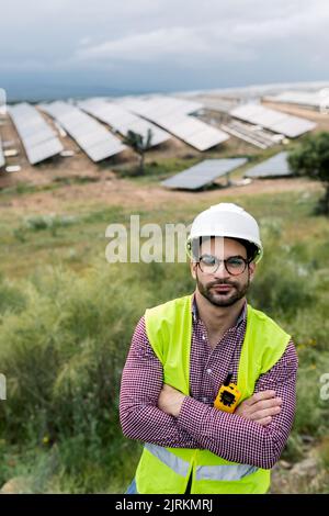 Felice ingegnere maschio con le braccia incrociate sorridendo guardando la macchina fotografica mentre si trova in piedi su sfondo sfocato di pannelli fotovoltaici e cielo nuvoloso Foto Stock