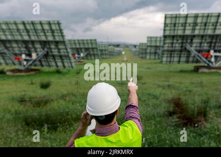 Vista posteriore dell'uomo anonimo in hardhat che indica i pannelli fotovoltaici mentre discute il lavoro della centrale di energia solare sullo smartphone Foto Stock