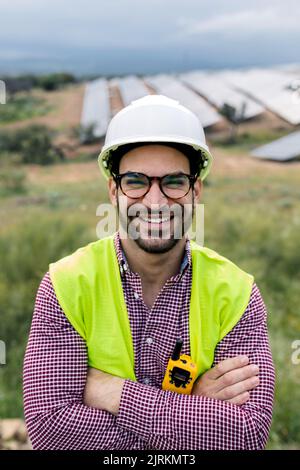 Felice ingegnere maschio con le braccia incrociate sorridendo guardando la macchina fotografica mentre si trova in piedi su sfondo sfocato di pannelli fotovoltaici e cielo nuvoloso Foto Stock