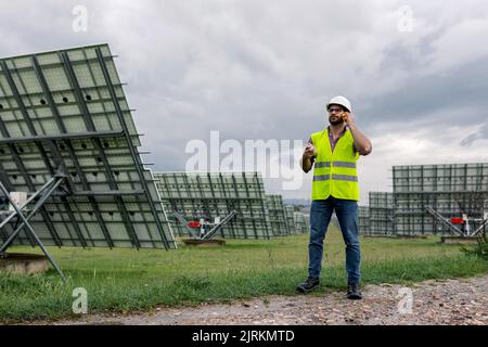 Maschio che tiene il BluePrint mentre parla sul walkie talkie contro la stazione di energia solare e il cielo grigio sovrastato Foto Stock