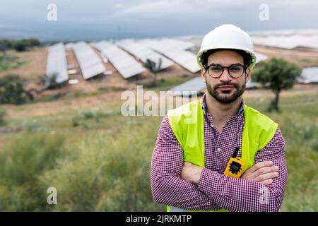 Felice ingegnere maschio con le braccia incrociate sorridendo guardando la macchina fotografica mentre si trova in piedi su sfondo sfocato di pannelli fotovoltaici e cielo nuvoloso Foto Stock