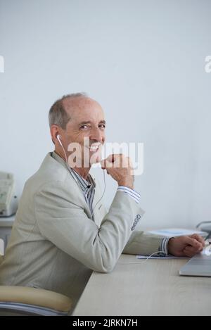 Uomo d'affari allegro e anziano in auricolari che lavorano al suo tavolo Foto Stock
