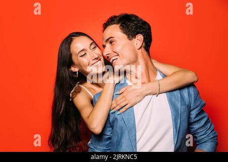 Giovane donna allegra che abbraccia fidanzato candido in abito elegante con taglio di capelli moderno mentre si guarda l'un l'altro Foto Stock