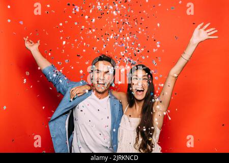 Donna allegra che abbraccia un uomo sorridente in un abbigliamento elegante mentre guarda la fotocamera con le braccia sollevate sotto gli streamer su sfondo rosso Foto Stock
