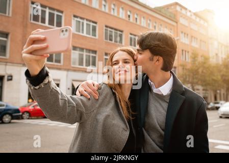 Felice giovane uomo che abbraccia e bacia la ragazza che porta selfie sullo smartphone mentre si trova in piedi insieme sulla strada della città Foto Stock