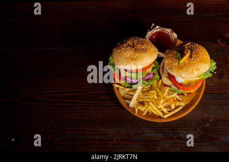 Due deliziosi hamburger fatti in casa di manzo con patatine fritte, formaggio e verdure su un vecchio tavolo di legno. Primo piano su cibi grassi e non sani Foto Stock