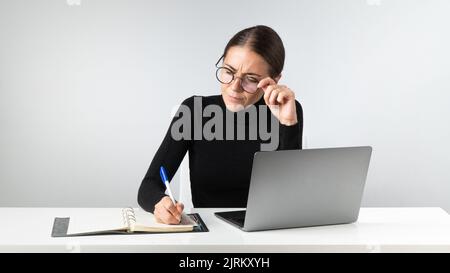 Una donna d'affari seria lavora in ufficio con un portatile, prende appunti in un notebook Foto Stock