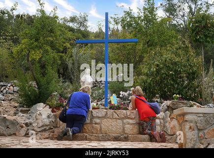 Due donne che si inginocchiano in preghiera ai piedi del Podbrdo (collina delle apparizioni), davanti alla croce blu e alla statua della Beata Vergine Maria (Medjugorje) Foto Stock