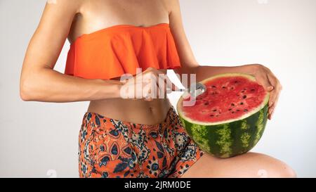 Una donna in costume da bagno mangia l'anguria con un cucchiaio su sfondo bianco - ora legale Foto Stock