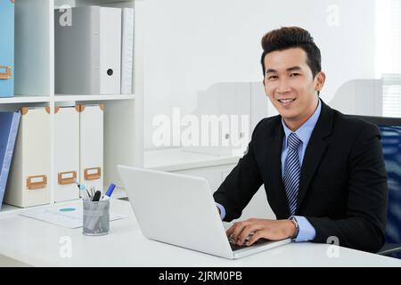 Bel colletto bianco asiatico indossando abito seduto di fronte al moderno computer portatile e posa per la fotografia, moderno ufficio interno sullo sfondo Foto Stock