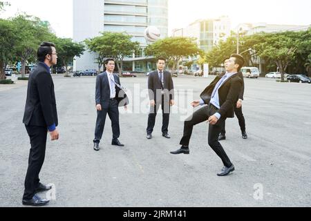 Cinque manager asiatici che indossano abiti classici che giocano a calcio all'aperto per fare poco esercizio fisico dopo una faticosa giornata di lavoro in ufficio Foto Stock