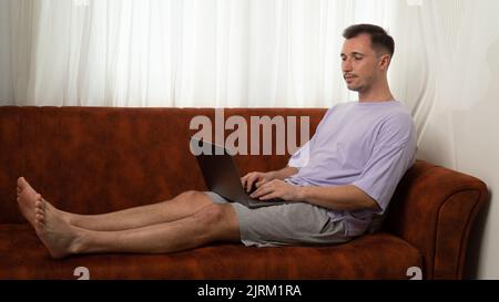Un uomo sdraiato sul divano e digita un laptop, lavorando al computer di casa Foto Stock