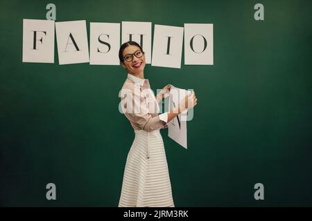 Designer femminile in piedi vicino alla parete e attaccando lettere di parole di moda. Giovane donna che tiene l'alfabeto per attaccare sulla parete dello studio. Foto Stock