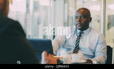 Uomo d'affari afroamericano in abbigliamento formale che spiega il suo piano di startup per partner in caffè vetroso durante l'ora di pranzo Foto Stock