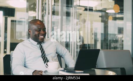 Ritratto di esilarante uomo d'affari afro-americano in abiti formali sorridendo, stampando e lavorando sul suo computer portatile in arioso caffè durante la pausa pranzo. Foto Stock