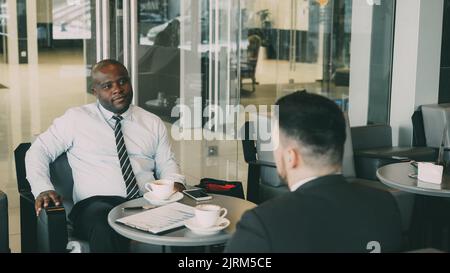 Sorridente uomo d'affari bere caffè e discutere rapporto finanziario di startup con il suo partner in moderno caffè Foto Stock