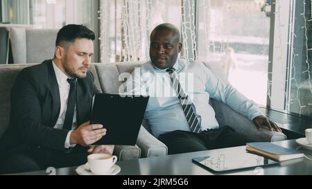 Due uomini d'affari multi-etnici che guardano gli appunti e discutono i progetti di startup in un moderno cafè durante l'ora di pranzo Foto Stock
