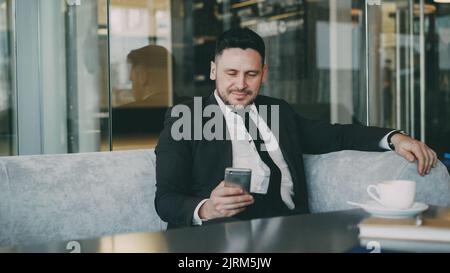 Sorridente uomo d'affari caucasico in abiti formali sorridente, rilassante e guardando lo schermo del suo smartphone mentre beve caffè in un arioso caffè durante l'ora di pranzo Foto Stock