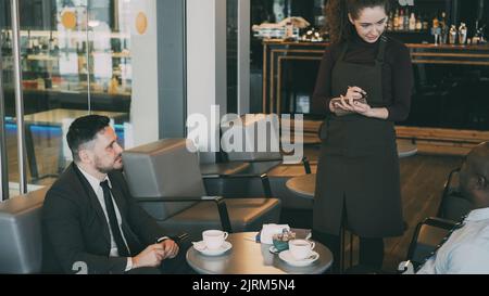 Due allegri uomini d'affari multietnici in abiti formali seduti, sorridenti e ordinanti in un moderno cafè durante la pausa pranzo. Cameriera giovane e ottimista che scrive i loro piatti Foto Stock