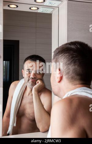 Un uomo guarda da vicino nello specchio del bagno, protendendo la guancia con la mano e guardando tristemente il viso. Foto Stock