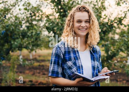 L'esperto agricolo sorridendo alla macchina fotografica felice che ha finito di esaminare gli alberi di prugne, tenendo un taccuino in cui la scrive Foto Stock