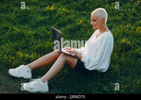 Ritratto di giovane millenaria donna europea dai capelli corti che usa un computer portatile sul prato verde nel parco. Bella ragazza bionda felice all'aperto. Abbigliamento estivo di moda femminile. Foto Stock