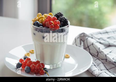 Yogurt, cereali d'avena per la colazione e frutti freschi d'estate in bicchiere su un tavolo bianco. Ribes rossi, mirtilli, more su piatto bianco, Foto Stock