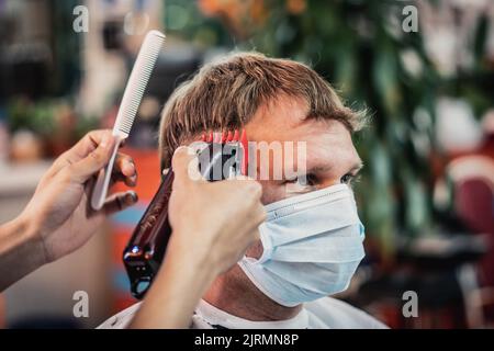 Foto stile di vita. Barbiere professionale. Il giovane cliente indossa una maschera facciale. Parrucchiere mani maschili Work Move pettine utensile regolacapelli elettrico. Moderno Foto Stock