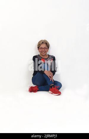 donna più anziana siede sul pavimento e si appoggia contro una parete su sfondo bianco Foto Stock