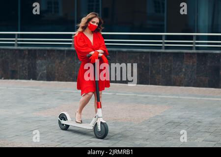 Giovane donna con scooter elettrico in abito rosso e guanti con maschera facciale in città. Nuovo concetto di protezione COVID per la moda normale e il coronavirus Foto Stock