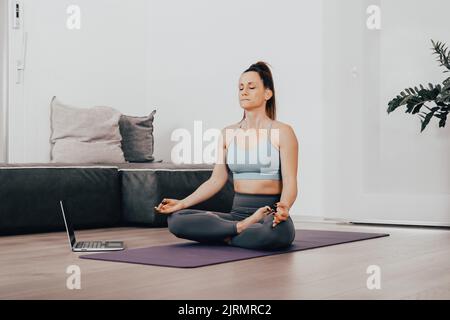 Donna calma vestita di abbigliamento sportivo, medita sul pavimento nel suo salotto, ascoltando lezioni di pratica spirituale sul computer portatile, posa in posa lotus, Foto Stock