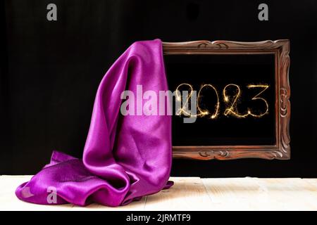 2023 numero scritto con scintillatori all'interno di una cornice marrone. Tessuto satinato viola che svela una cornice in legno vintage su sfondo nero. Felice anno nuovo mock Foto Stock