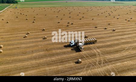 Il trattore agricolo carica la paglia di frumento sul dumper. Vista aerea. Macchine agricole. Cataste di fieno dopo la raccolta di prodotti di grano. Foto Stock