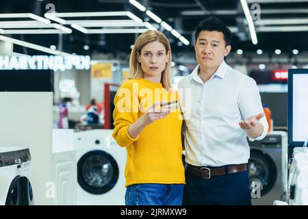 Frustrata giovane famiglia diversificata di acquirenti, uomo e donna che guarda la macchina fotografica, non avendo abbastanza soldi sulla carta di credito per acquistare beni elettronici in supermercato Foto Stock