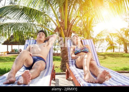 Giovane coppia vietnamita che prende il sole sulle chaise-lounge sotto la palma Foto Stock