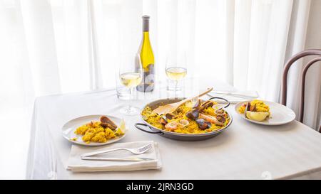 Tavolo del ristorante con piatto tradizionale spagnolo. Paella di pesce con gamberi, vongole, cozze su riso allo zafferano e verdure e bottiglia di vino. Foto Stock