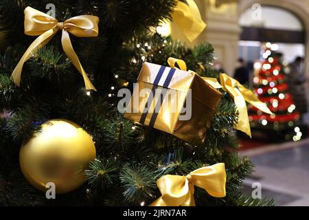 Albero di Natale con giocattoli dorati e scatola regalo in un centro commerciale sullo sfondo di luci festive sfocate. Decorazioni di Capodanno, vacanze invernali Foto Stock