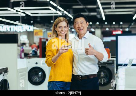 Ritratto di famiglia diverse coppie asiatico uomo e donna shopping in supermercato, elettronica e elettrodomestici, guardando la macchina fotografica e sorridente, soddisfatto con carta di credito bancario Foto Stock