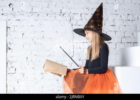 Halloween bambini. Una bella dolce ragazza in costume da strega, indossando un cappello, tenendo un libro con incantesimi e una bacchetta magica, congiura seduto sui gradini. Foto Stock