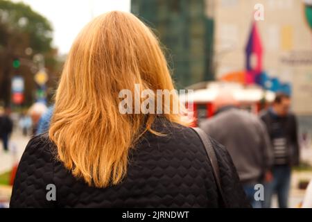 Defocalizzare il concetto di cittadino urbano. Vista posteriore della donna bionda che attraversa la strada in città, con pendolari ed edifici commerciali sullo sfondo. Primo piano uno p Foto Stock